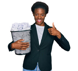 Sticker - Young african american girl holding paper bin full of crumpled papers smiling happy and positive, thumb up doing excellent and approval sign