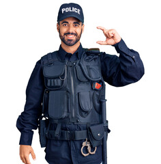 Poster - Young hispanic man wearing police uniform smiling and confident gesturing with hand doing small size sign with fingers looking and the camera. measure concept.