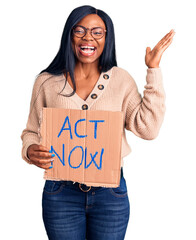 Wall Mural - Young african american woman holding act now banner celebrating victory with happy smile and winner expression with raised hands