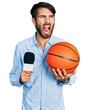 Hispanic man with blue eyes holding reporter microphone and basketball ball angry and mad screaming frustrated and furious, shouting with anger. rage and aggressive concept.
