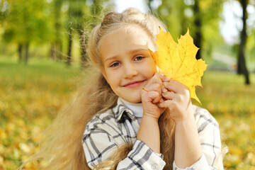 Wall Mural - A little preschool blonde girl holds an autumn leaf in her hands, smiles and looks at the camera