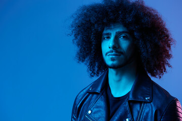 Portrait of fashion man with curly hair on blue background multinational, colored light, black leather jacket trend, modern concept.