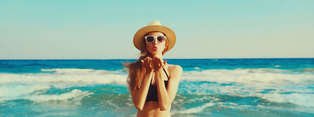 Summer vacation, beautiful happy woman blowing her lips sends air kiss in bikini swimsuit and straw hat on the beach on sea coast with waves background