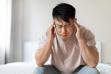 Sad middle aged asian man sitting on bed, touching head