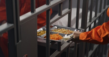 Elderly prisoner in orange uniform sits in prison cell. Prison guard gives him tray of food through metal bars. Guilty criminal serves imprisonment term for crime. Inmate in jail or detention center.