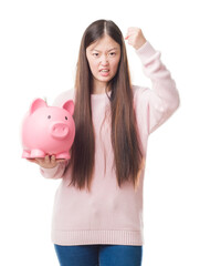 Canvas Print - Young Chinese woman over isolated background holding piggy bank annoyed and frustrated shouting with anger, crazy and yelling with raised hand, anger concept