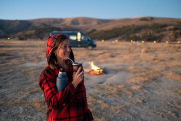 Wall Mural - A woman drinking traditional Argentine mate by a campfire at dusk. Behind her campervan