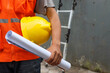 Front view of male construction worker holding blueprint and safety helmet outdoor of construction site with ladder in the background. Copy space and selective focus.
