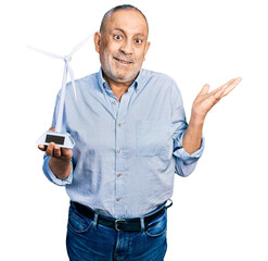 Canvas Print - Senior man with grey hair and beard holding solar windmill for renewable electricity celebrating victory with happy smile and winner expression with raised hands