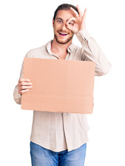 Sticker - Young handsome caucasian man holding empty cardboard banner smiling happy doing ok sign with hand on eye looking through fingers