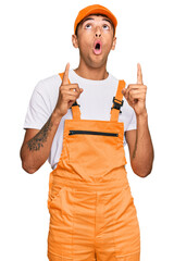 Canvas Print - Young handsome african american man wearing handyman uniform amazed and surprised looking up and pointing with fingers and raised arms.