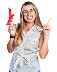 Wall Mural - Hispanic young woman holding graduate degree diploma smiling happy pointing with hand and finger to the side