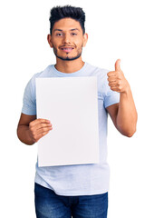 Wall Mural - Handsome latin american young man holding cardboard banner with blank space smiling happy and positive, thumb up doing excellent and approval sign