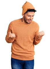 Sticker - Handsome latin american young man wearing winter sweater and wool hat very happy and excited doing winner gesture with arms raised, smiling and screaming for success. celebration concept.