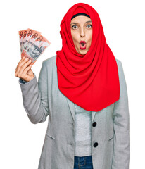 Sticker - Beautiful hispanic woman wearing islamic hijab holding australian dollars banknotes scared and amazed with open mouth for surprise, disbelief face