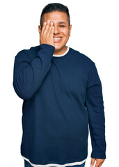 Poster - Young latin man wearing casual clothes covering one eye with hand, confident smile on face and surprise emotion.