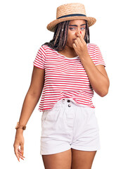 Canvas Print - Young african american woman with braids wearing summer hat smelling something stinky and disgusting, intolerable smell, holding breath with fingers on nose. bad smell