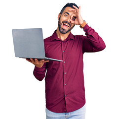 Wall Mural - Young hispanic man holding laptop smiling happy doing ok sign with hand on eye looking through fingers