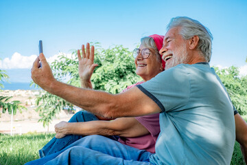 Wall Mural - Video call concept. Happy senior couple sitting in the park using smartphone for online communication via video meeting, smiling grey-haired man and woman looking and talking into phone webcam