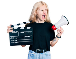 Canvas Print - Young caucasian woman holding video film clapboard and megaphone angry and mad screaming frustrated and furious, shouting with anger. rage and aggressive concept.