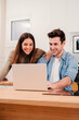 © Jose Calsina - Vertical portrait of young couple browsing on internet at home kitchen using a laptop. Happy husband and wife planning the vacations with a computer talking and smiling together, watching funny videos