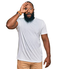 Poster - Young african american man wearing casual white tshirt doing ok gesture shocked with surprised face, eye looking through fingers. unbelieving expression.