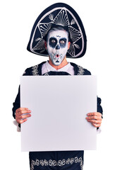 Wall Mural - Young man wearing day of the dead costume holding blank empty banner scared and amazed with open mouth for surprise, disbelief face