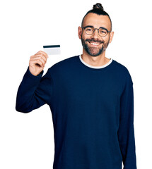 Hispanic man with ponytail holding credit card looking positive and happy standing and smiling with a confident smile showing teeth