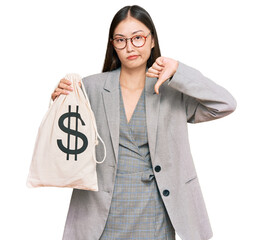 Sticker - Young chinese woman wearing business suit holding dollars bag with angry face, negative sign showing dislike with thumbs down, rejection concept