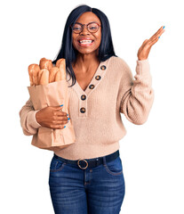 Poster - Young african american woman holding paper bag with bread celebrating victory with happy smile and winner expression with raised hands