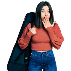 Canvas Print - Young brunette woman with blue eyes wearing guitar case covering mouth with hand, shocked and afraid for mistake. surprised expression