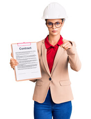 Sticker - Young beautiful woman wearing architect hardhat holding clipboard with contract document pointing with finger to the camera and to you, confident gesture looking serious