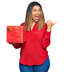 Poster - Beautiful brunette woman holding gift pointing thumb up to the side smiling happy with open mouth