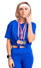 Wall Mural - Young blonde woman wearing winner medals serious face thinking about question with hand on chin, thoughtful about confusing idea