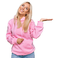 Poster - Young blonde woman wearing casual sweatshirt smiling cheerful presenting and pointing with palm of hand looking at the camera.