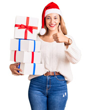 Beautiful Young Woman Wearing Christmas Hat And Holding A Gift Smiling Happy And Positive, Thumb Up Doing Excellent And Approval Sign
