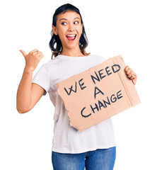 Sticker - Young woman holding we need a change banner pointing thumb up to the side smiling happy with open mouth