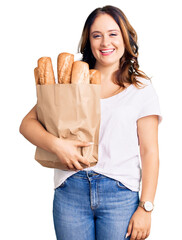 Young beautiful caucasian woman holding paper bag with bread looking positive and happy standing and smiling with a confident smile showing teeth