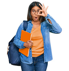 Canvas Print - Young latin girl wearing student backpack and holding books doing ok gesture shocked with surprised face, eye looking through fingers. unbelieving expression.