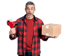 Poster - Middle age hispanic with grey hair holding packing tape and cardboard box afraid and shocked with surprise and amazed expression, fear and excited face.