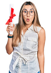 Canvas Print - Hispanic young woman holding graduate degree diploma scared and amazed with open mouth for surprise, disbelief face