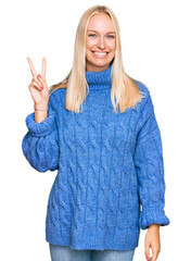 Wall Mural - Young blonde girl wearing wool winter sweater smiling with happy face winking at the camera doing victory sign. number two.