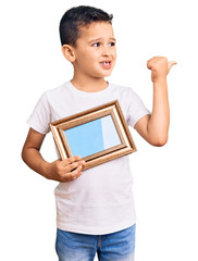 Wall Mural - Little cute boy kid holding empty frame pointing thumb up to the side smiling happy with open mouth