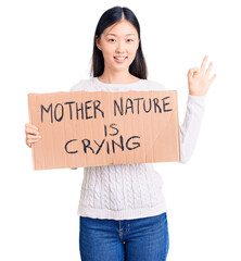 Poster - Young beautiful chinese woman holding mother nature is crying cardboard banner doing ok sign with fingers, smiling friendly gesturing excellent symbol