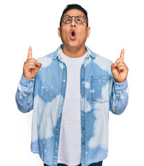 Sticker - Young latin man wearing casual clothes and glasses amazed and surprised looking up and pointing with fingers and raised arms.