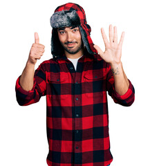 Wall Mural - Hispanic young man with beard wearing fluffy earmuff hat showing and pointing up with fingers number six while smiling confident and happy.