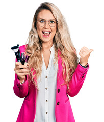 Poster - Young blonde woman holding makeup brushes pointing thumb up to the side smiling happy with open mouth