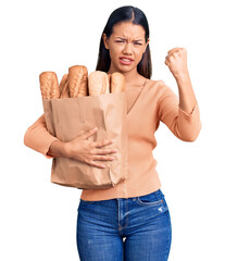 Poster - Young beautiful latin girl holding paper bag with bread annoyed and frustrated shouting with anger, yelling crazy with anger and hand raised