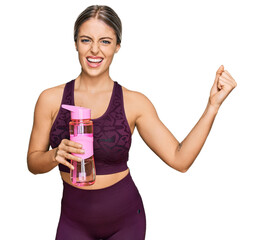 Wall Mural - Beautiful blonde woman wearing sportswear holding water bottle screaming proud, celebrating victory and success very excited with raised arms