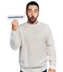 Wall Mural - Young hispanic man with beard holding ransomware banner scared and amazed with open mouth for surprise, disbelief face